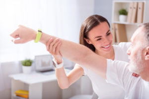 A senior man in a physical therapy session working with a smiling therapist who is helping him extend his left arm to shoulder height.